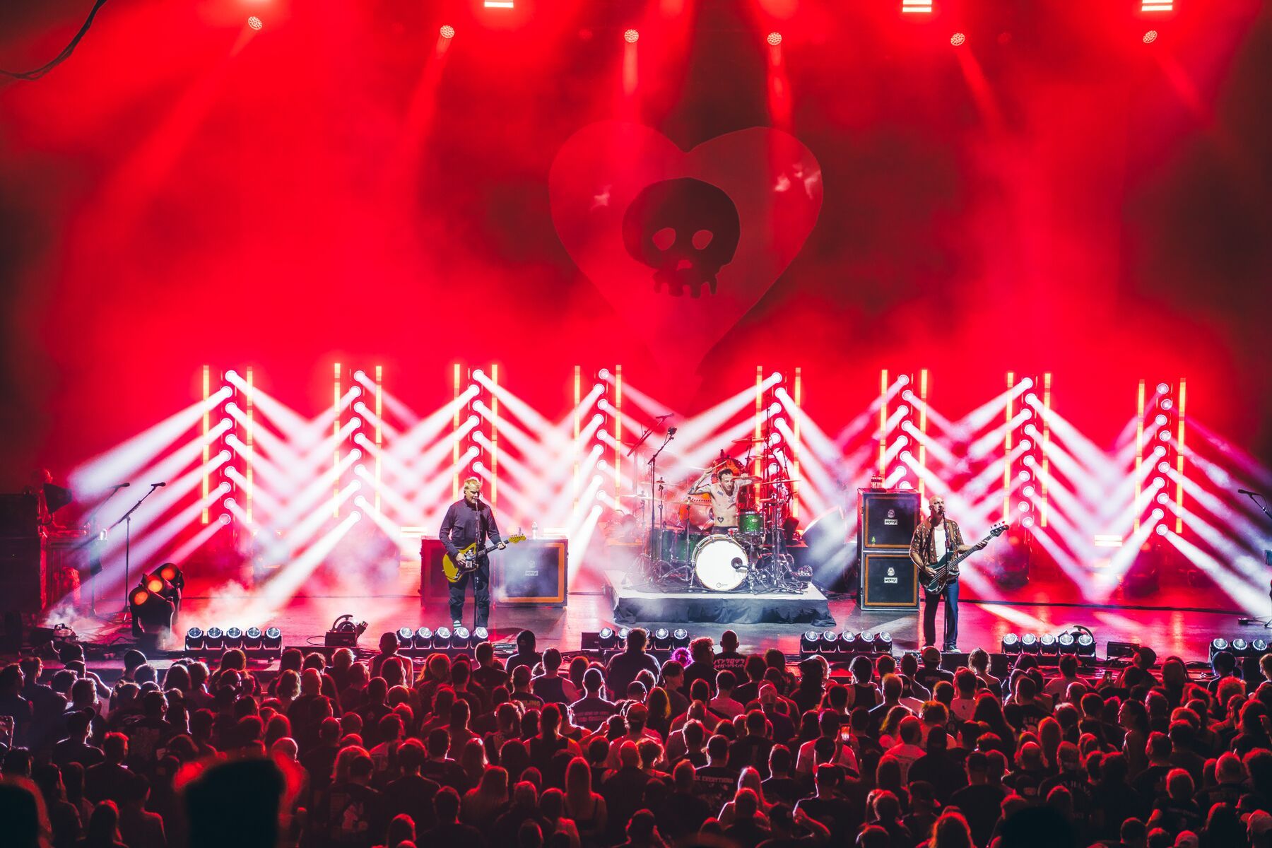 Alkaline Trio on stage under red light, with white chevron light fixtures across the rig